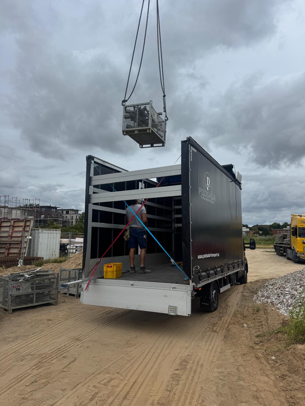 Crane loading a metal crate onto a Podsada Transport trailer – construction site
